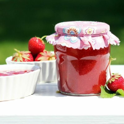 Fresh homemade strawberry jam in a decorative jar with ceramic bowls on a white table outdoors.