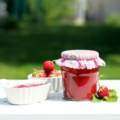 Fresh homemade strawberry jam in a decorative jar with ceramic bowls on a white table outdoors.